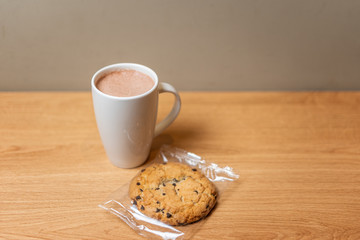 A mug of cocoa on a wooden table next to an oatmeal cookie.