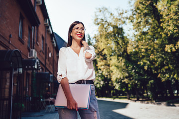 Fototapeta premium Cheerful charming woman in classic eyewear holding laptop for e learning and smiling at city urban setting, young millennial female student trendy dressed looking away and feeling good outdoors