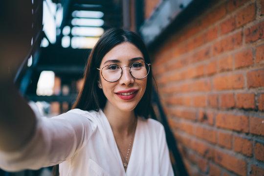 Close up portrait of charming Spanish hipster girl with cute smile on face making selfie images during leisure time at urban setting, beautiful woman photographing herself posing at city street
