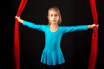 Fearless pretty little girl in a blue gymnastic suit shows a stunt aerial red ribbon on a black...