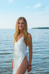 Fototapeta na wymiar Beautiful young woman in a white summer satin dress stands at sea background