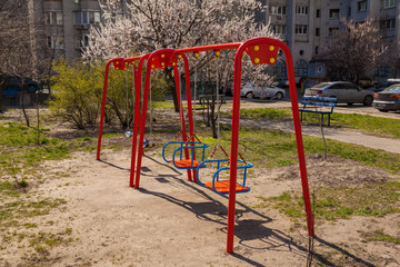Fototapeta na wymiar Empty swing on the playground during quarantine due to coronavirus on the background of a blossoming white tree and a multi-storey residential building