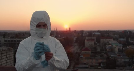 Portrait of a young male in protective suit and respiratry mask removing his gloves while standing on a roof with a cityscape view in a sunny spring sunset during pandemic coronavirus quarantine time - Powered by Adobe