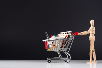 Wooden doll rolls metal shopping cart with pills on dark background. Pills, tablets and medicine on black background. Healthcare concept.