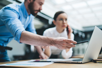 Adult businessman pointing on laptop screen while colleague phoning at desk in office