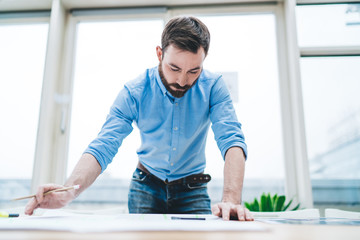 Fototapeta na wymiar Adult man intently working with blueprint while standing at desk in open space office