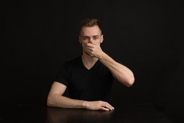 a man in a black shirt on a black background covers his mouth with his hand