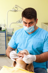 Dentist examining patient's teeth, wearing blue uniform and gloves, looking at patient's teeth carefully with dental instruments