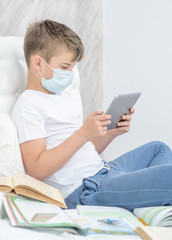 Boy with books wearing medical mask uses tablet computer at home. Quarantine and coronavirus epidemic concept
