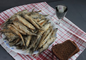 Smelt fried on a plate and dish towel. Next to a few slices of bread and a glass with vodka. Dark background. Close up. View from above.