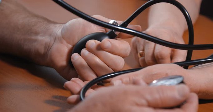 Close-up detail shot of hands of an elderly couple. Husband takes care of his wife by measuring her blood pressure using a mechanical tonometer on a brown wooden table at home. 4k 50fps slow motion