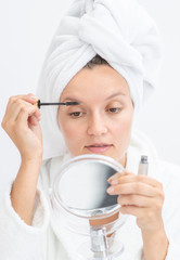 Young woman holds mirror and applies makeup at home