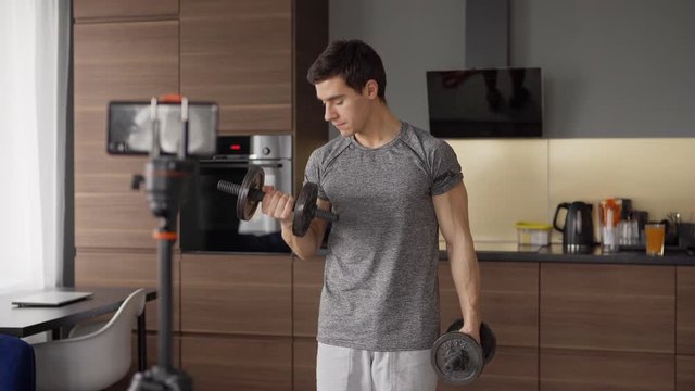 Tilt Up Medium Shot Of Young Male Fitness Blogger Recording Dumbbell Bicep Curl Exercise On Cell Phone Placed On Tripod At Home