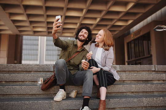 Cheerful young couple in formal clothing taking selfie using smartphone while eating sandwich for lunch during break