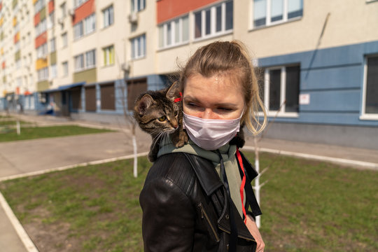 a girl in a medical mask is walking with a cat along the street near the house