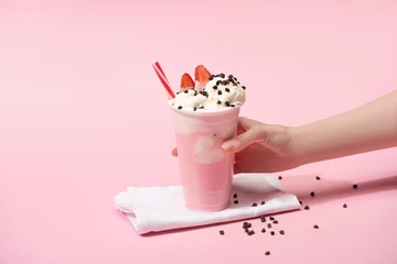 Tuinposter Cropped view of female hand with disposable cup of milkshake with chocolate morsels and strawberry on napkins on pink © LIGHTFIELD STUDIOS