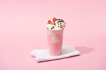 Disposable cup of milkshake with chocolate chips and strawberry halves on napkins on pink background
