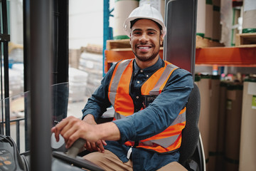 Portrait of happy driver wearing white helmet and vest sitting in forklift machine transporting...