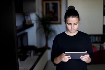 Adolescente  con i capelli scuri raccolti e un maglia nera guarda il tablet in salotto 