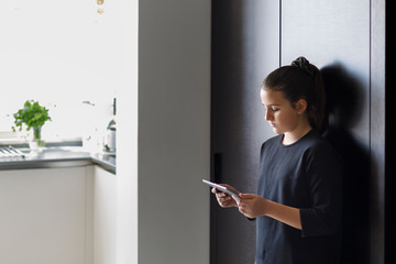 Adolescente  con i capelli scuri raccolti e un maglia nera guarda il tablet in salotto 