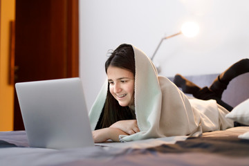 Giovane ragazza coni capelli scuri chatta sorridente al computer a letto con un a coperta in testa
