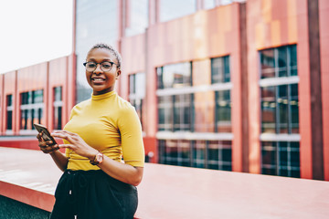 Naklejka premium Half length portrait of cheerful woman in classic glasses for eyes correction holding cellphone technology and smiling at camera, African American female blogger with mobile gadget posing at urbanity