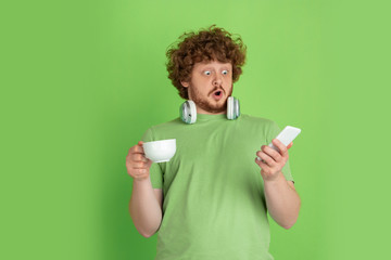 Enjoying coffee, surprised with smartphone. Caucasian young man's monochrome portrait on green background. Beautiful curly model in casual style. Concept of human emotions, facial expression, sales