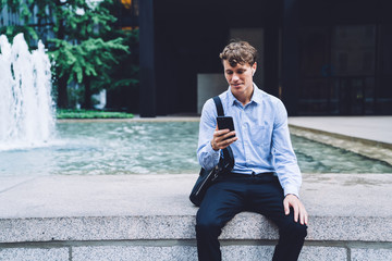 Smiling male with earbuds sitting at fountain and messaging on mobile on Manhattan