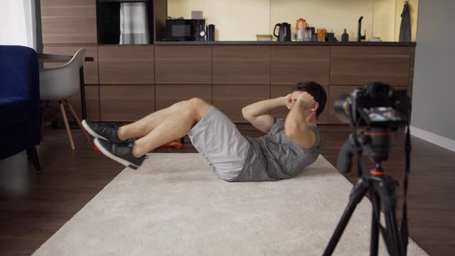 Young male fitness instructor performing abs cross crunches exercise lying on floor at home during livestream on cell phone placed on tripod