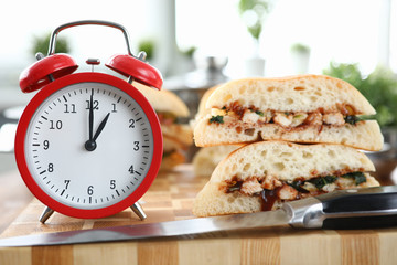 Close-up of red clock standing on wooden table and showing one pm on dial. Sandwiches and sharp knife. Lunch time. Break from work. Fast food delivery and nutrition concept