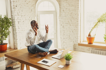 Young african-american man doing yoga at home while being quarantine and freelance online working. Remote, isolated or alone at office. Concept of healthy lifestyle, wellness, activity, movement.