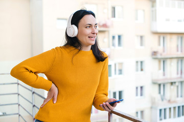 Woman enjoying morning time at balcony. Woman using mobile phone and wireless headphones. Self isolation at home.