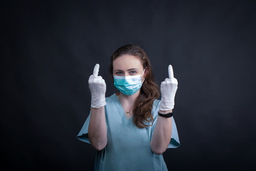 Female  nurse  wearing a turquoise surgical mask from coronavirus, double rude gesture, dark background