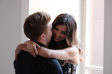 A young couple - a girl in a magnificent dress and a guy in a black shirt and trousers hug and kiss in a hotel room. 