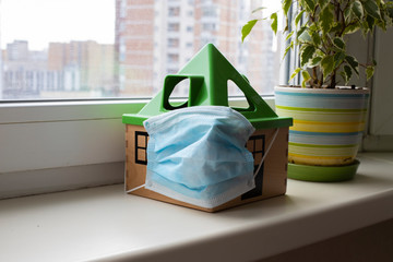 covid-19 pandemic, quarantine coronavirus. a toy house in a medical mask stands on a windowsill next to a flower in a pot.