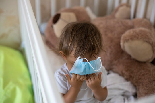 The Concept Of Quarantine And Self-isolation In The Pandemic Of The Coronavirus Covid -19. Little Cute Kid Hugs A Medical Mask While Sitting In A Crib At Home On The Background Of A Toy Bear