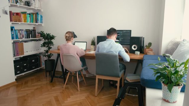 Couple or Partners Work in Home Office at Computer and Laptop at Daytime. Video Editor, Bloggers or Freelancers Work Remotely in Cozy Room. Family or Spouses Work Together. 4K Wide Static Shot