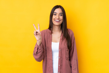 Young brunette woman over isolated yellow background smiling and showing victory sign