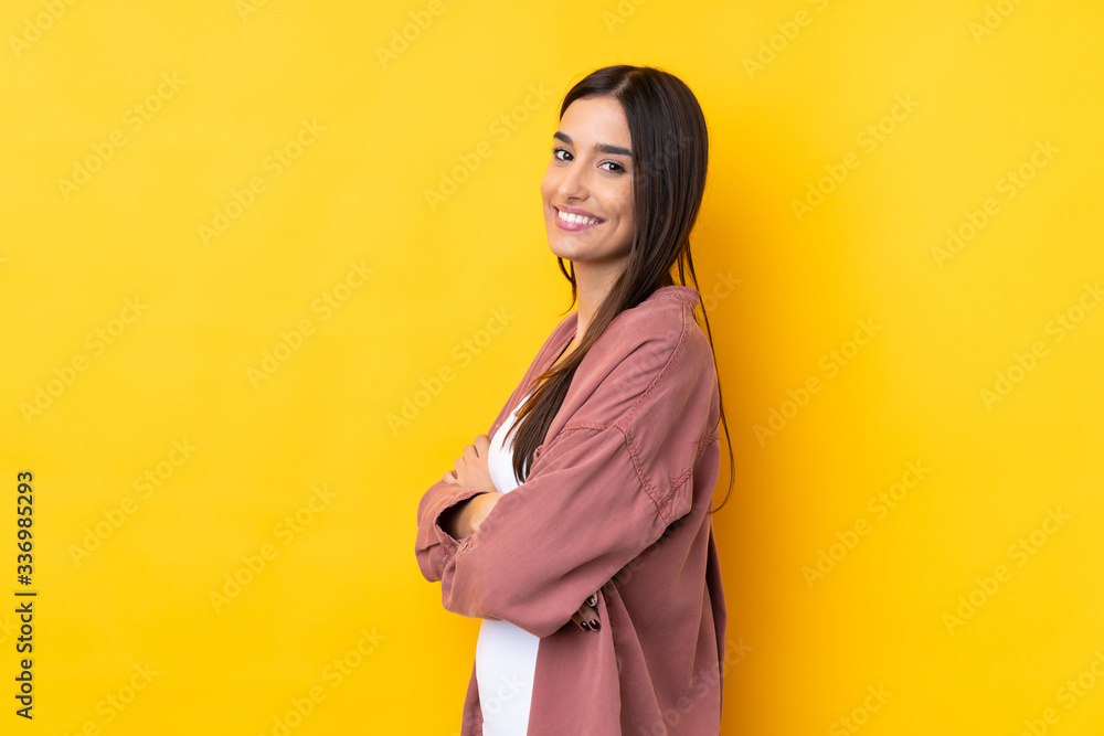 Wall mural young brunette woman over isolated yellow background with arms crossed and looking forward