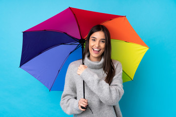 Young brunette woman holding an umbrella over isolated blue wall celebrating a victory