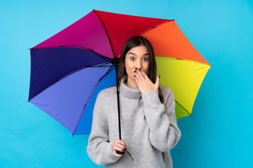 Young brunette woman holding an umbrella over isolated blue wall with surprise facial expression