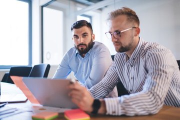Skilled bearded male boss looking at camera while his colleague share report about finance revenue of business, caucasian 20s men colleagues cooperating on common startup on working process in office