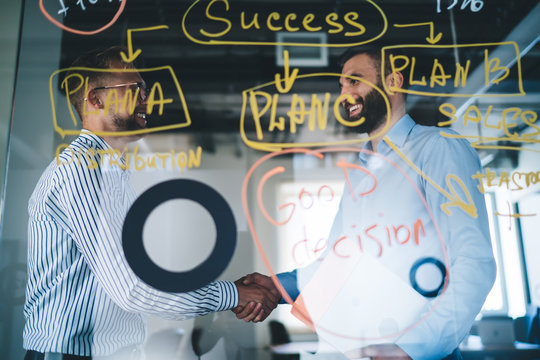 Smiling caucasian male employees shaking hands with new colleague on meeting standing near notes and graphic on glass, prosperous business partners gesturing agreement in productive negotiation - Powered by Adobe
