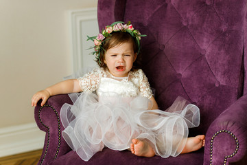 Little girl in a pink dress sits on a soft chair. Crying baby