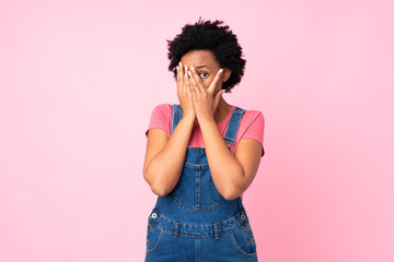 African american woman with overalls over isolated pink background covering eyes and looking through fingers
