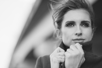 A beautiful lonely woman in a coat and scarf under a frozen bridge holds her collar with her hands....