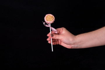 Cake pops in women's hands on a black background. Dessert in pink chocolate cream with powder and bow.