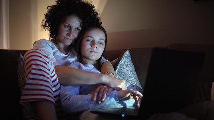 Mother and daughter in pajamas sitting on sofa doing online lessons during distance work and...