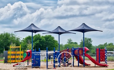 A public school playground sits deserted during the coronavirus quarantine period in Houston, TX. Three shade umbrellas stand out from the colorful equipment against a dramatic sky.