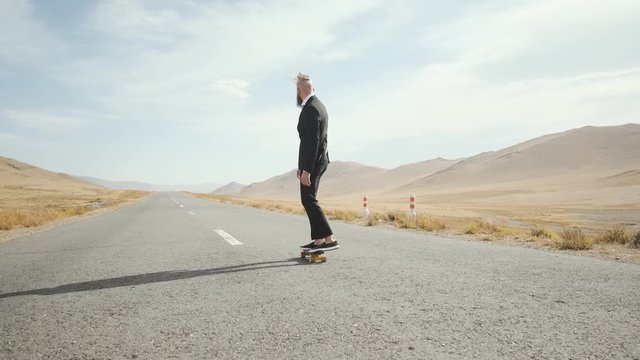 Caucasian man with beard and mustache in black suit and white shirt,an office worker happy excited man rides skateboard on an empty picturesque mountain road and drives away